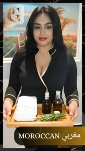Moroccan female spa therapist wearing a black uniform with gold trim, holding a wooden tray with white towels, oil bottles, green leaves, and a white flower at Golden Massage Center, Prime Hotel, 1st floor, Nakheel, Ras Al Khaimah. Background shows a peaceful beach-themed wall painting, highlighting spa ambience.