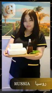 A young Russian female therapist in a black spa uniform with yellow trim, standing indoors at Golden Massage Center, Prime Hotel, Nakheel, Ras Al Khaimah. She holds a wooden tray with stacked white towels, a dark oil bottle with a pump top, and fresh green leaves. Behind her is a scenic beach wall painting, creating a relaxing spa environment. The words "RUSSIA" in English and Arabic appear on the image.